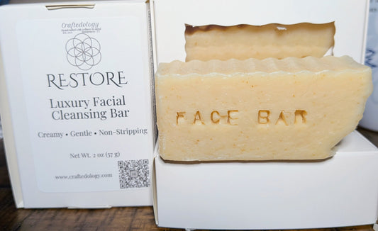A creamy off-white facial cleansing bar soap embossed with 'FACE BAR' sits in a white box next to its white packaging label for 'RESTORE Luxury Facial Cleansing Bar' by Craftedology.