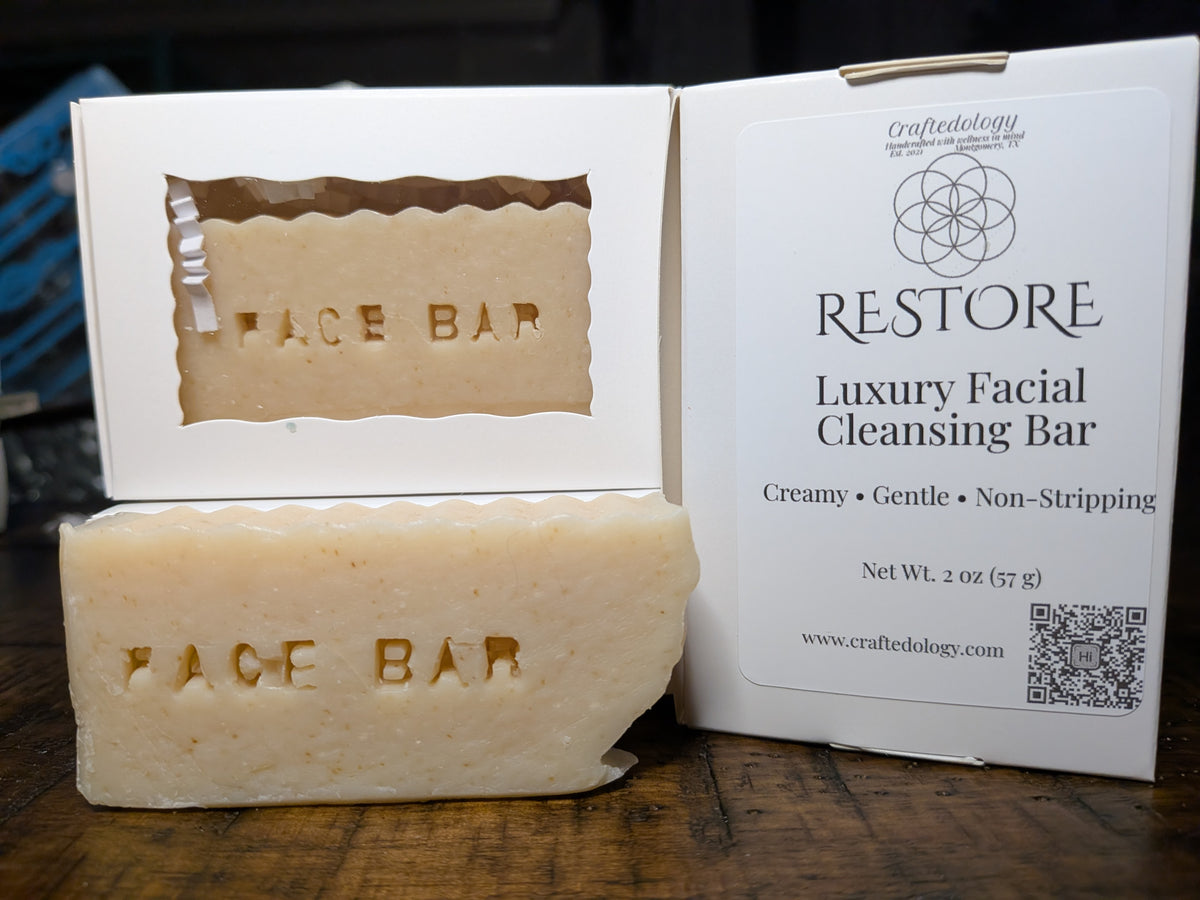A cream-colored facial cleansing bar soap with 'FACE BAR' embossed on it, shown next to its white packaging box labeled 'RESTORE Luxury Facial Cleansing Bar' by Craftedology, set on a dark wooden surface.